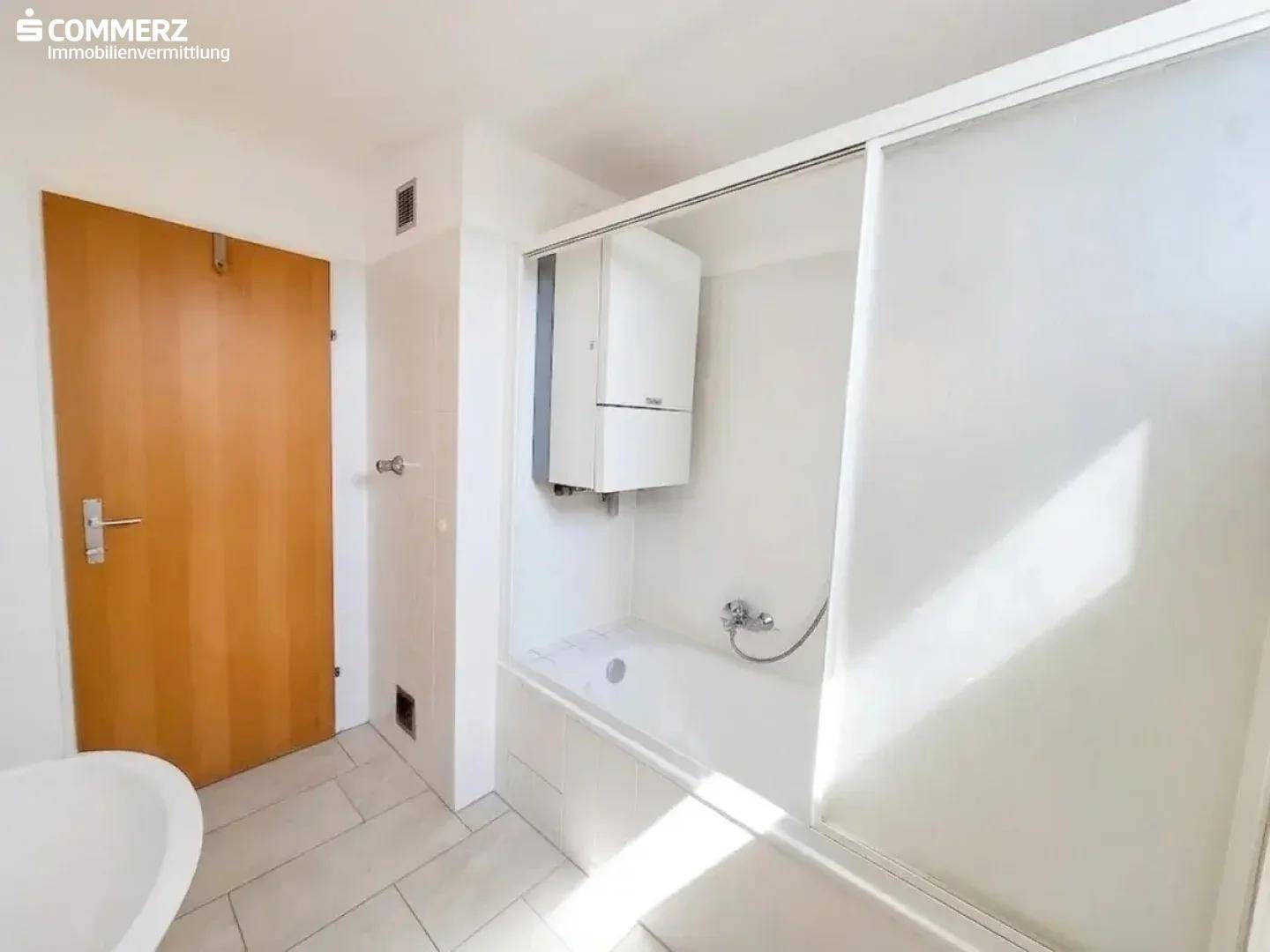 Helles Badezimmer mit Badewanne, Duschabtrennung, Heizung und Holztür.