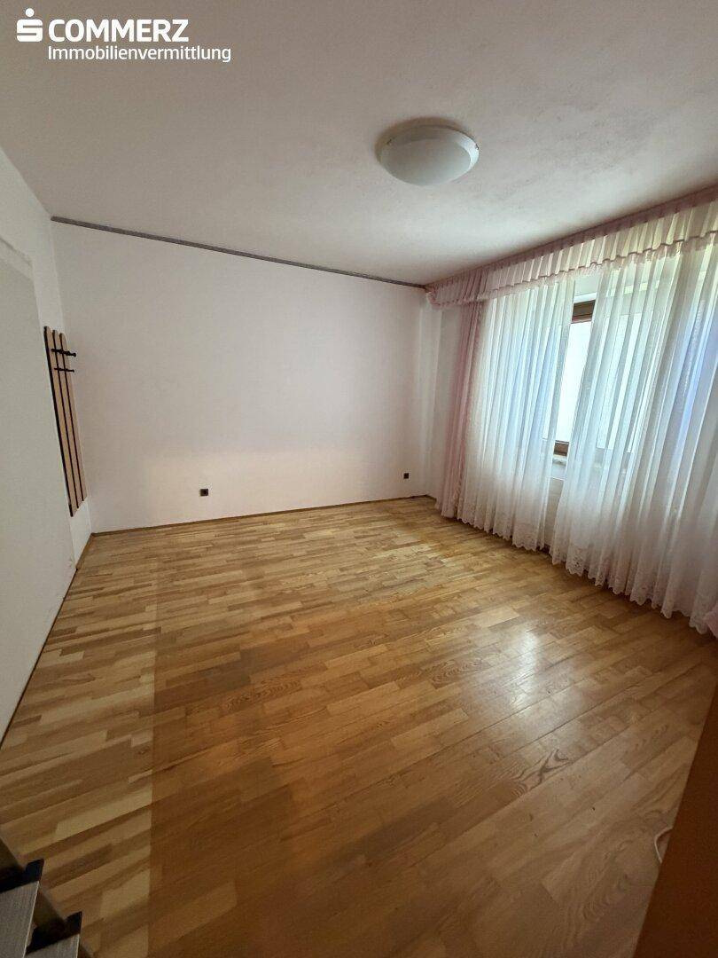 Schlafzimmer mit hellem Parkettboden, weißen Wänden und großem Fenster mit Vorhängen.