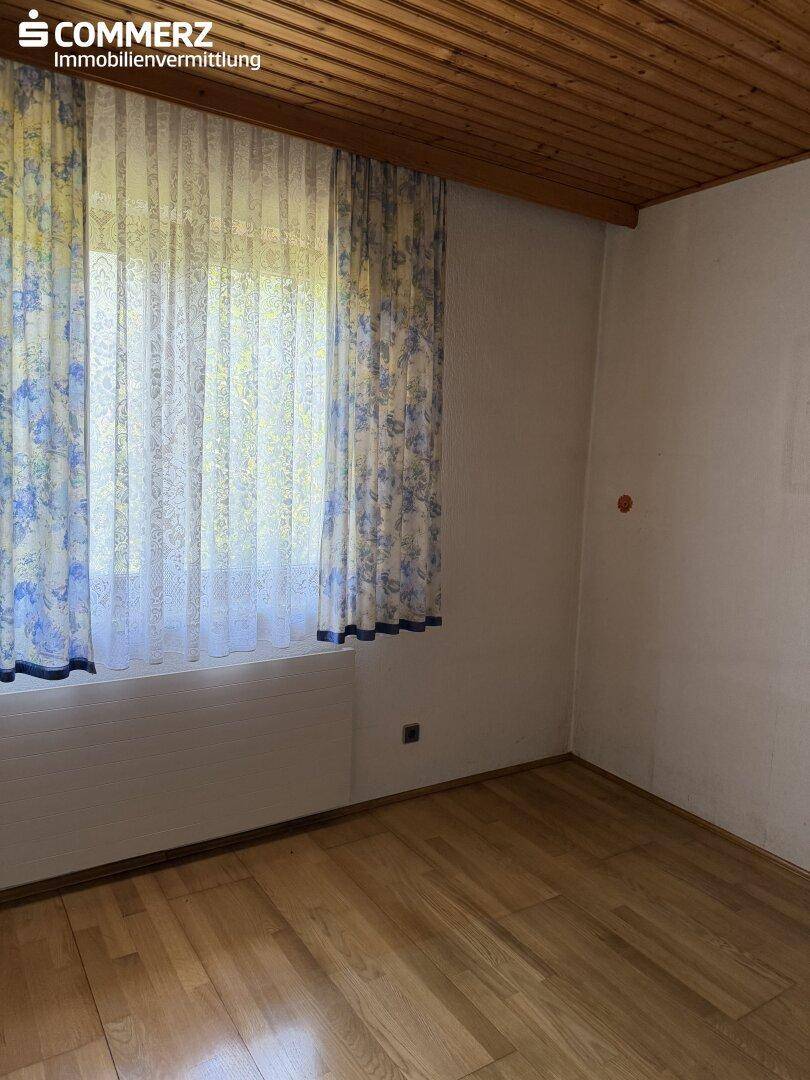 Schlafzimmer mit Holzdecke, Parkettboden und Fenster mit floralen Vorhängen.