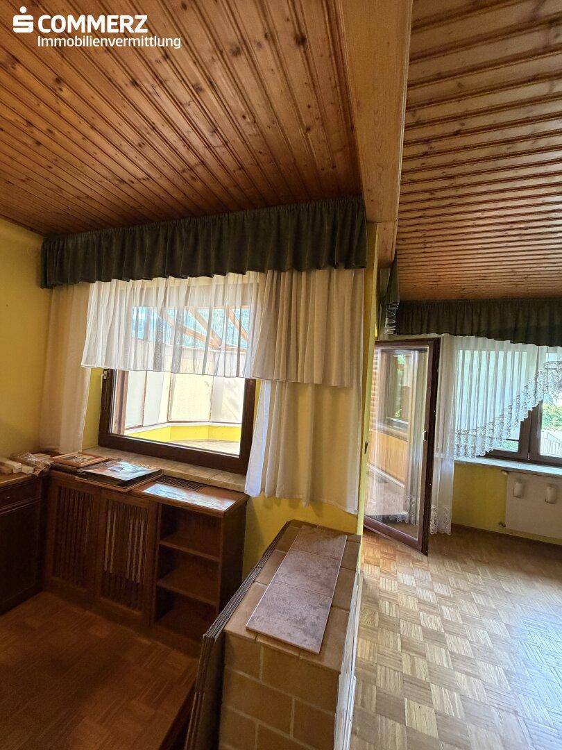 Wohnzimmerdetail mit Holzdecke, Parkettboden und Fenster mit Vorhängen.