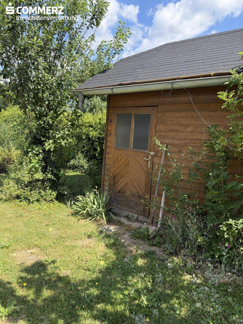 Kleiner Holzschuppen im Garten, umgeben von Bäumen und Sträuchern.
