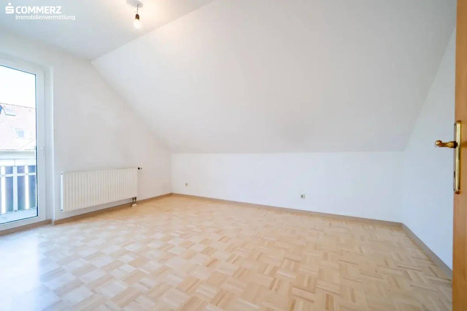 Helles Dachgeschosszimmer mit Parkettboden, Dachschräge und Zugang zu einem französischen Balkon.