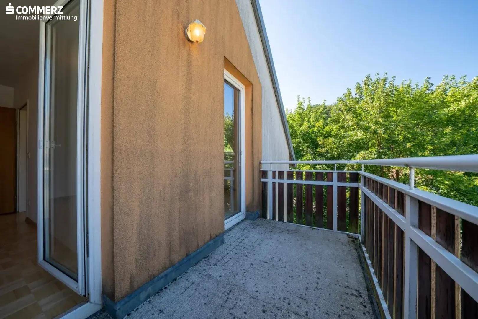 Kleiner Balkon mit Zugangstür und Blick auf die grüne Umgebung unter blauem Himmel.
