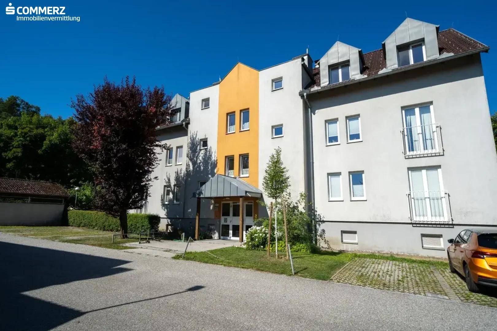 Mehrfamilienhaus mit heller Fassade, orangefarbenem Mittelteil und gepflegter Außenanlage.