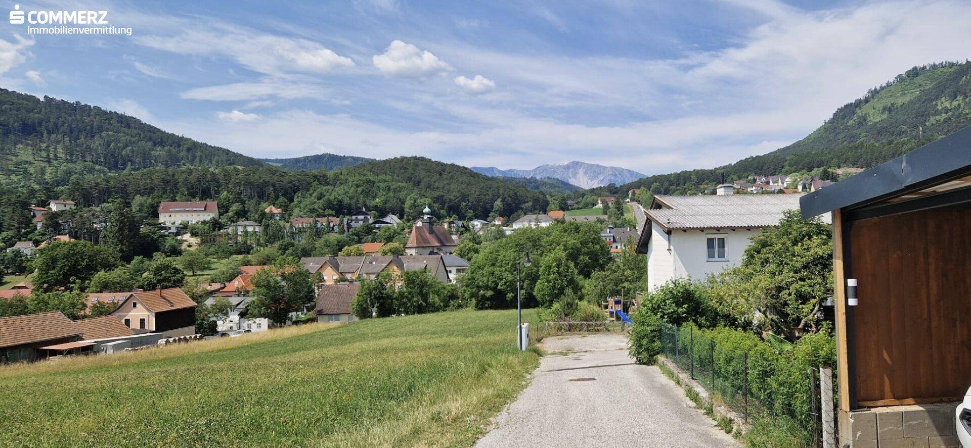 Weitläufiger Blick über eine ländliche Siedlung mit grünen Hügeln und Bergen im Hintergrund.