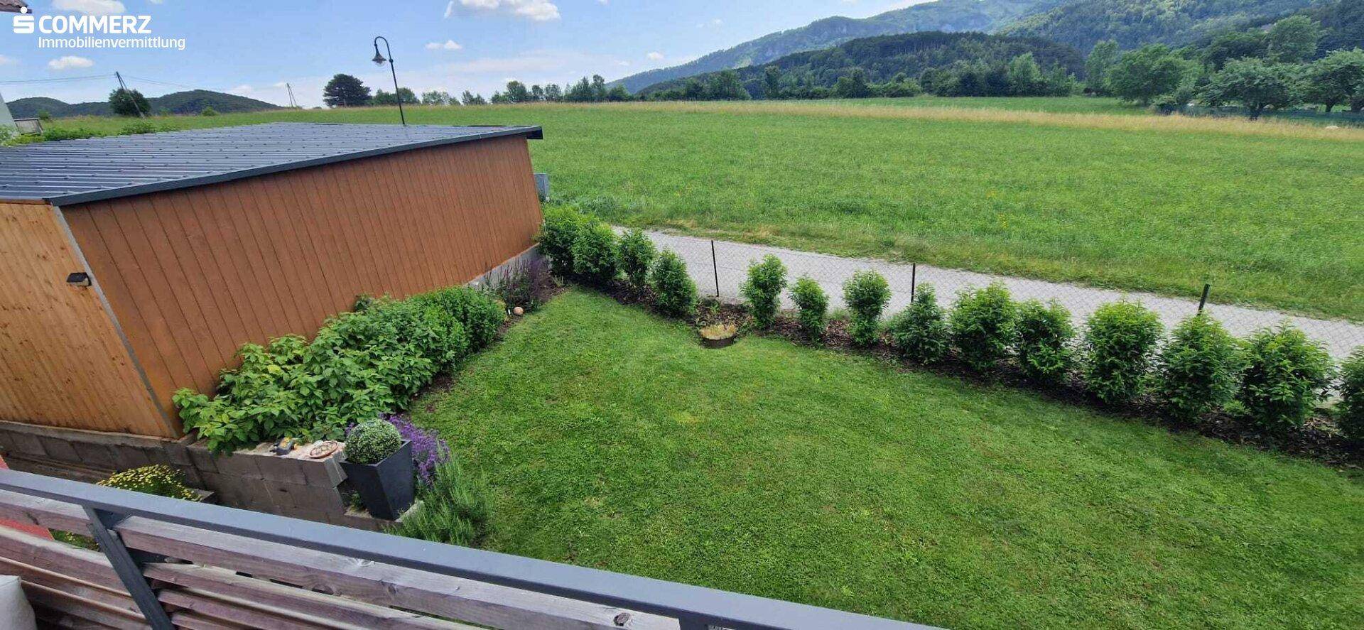 Gepflegter Rasenbereich mit Hecke und einem Holzschuppen, umgeben von grünen Feldern.