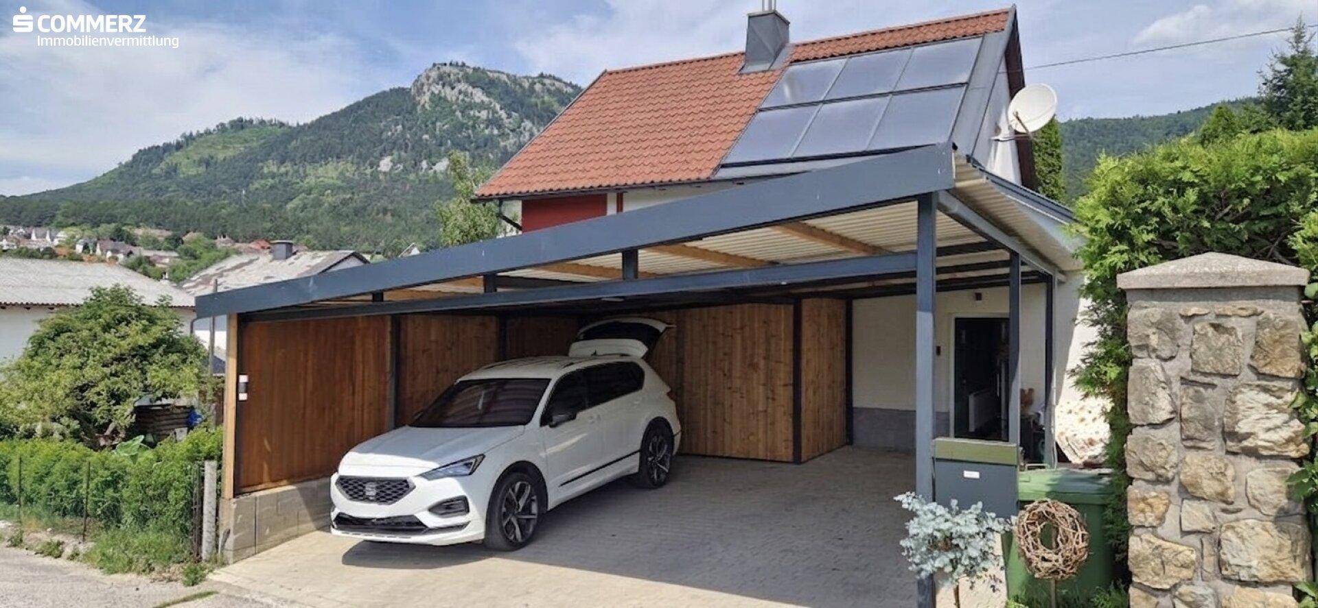 Einfamilienhaus mit Carport und Photovoltaikanlage auf dem Dach, umgeben von Bergen.