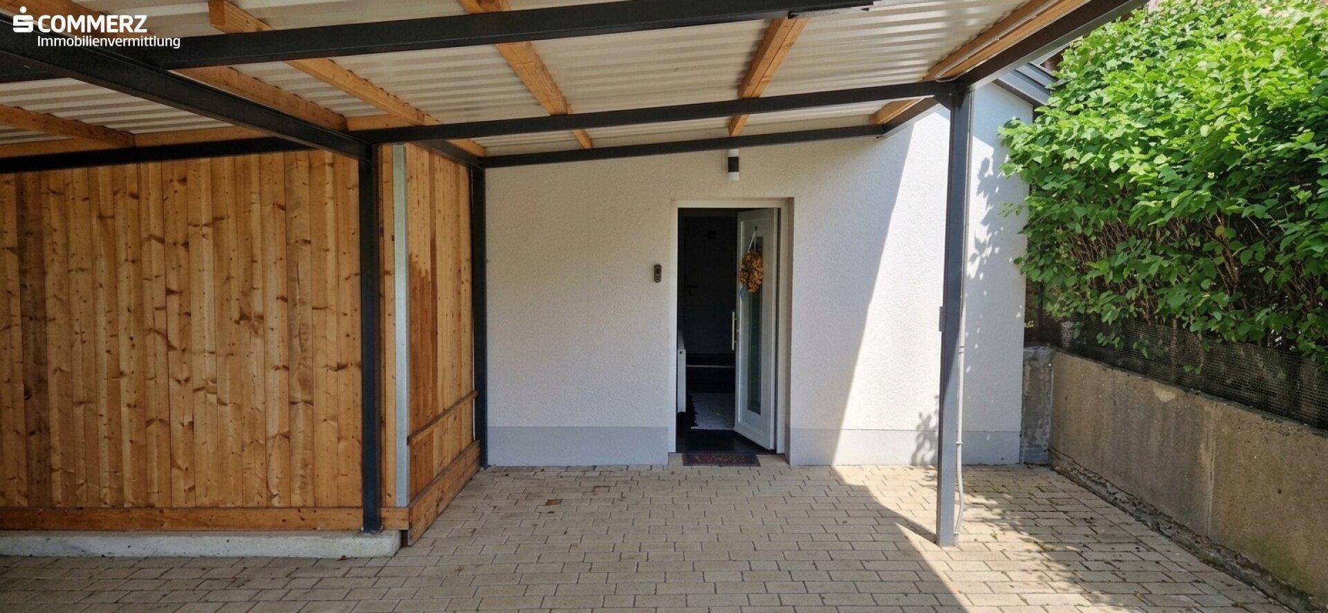 Überdachter Eingangsbereich unter einem Carport mit Holzwänden und gepflastertem Boden.