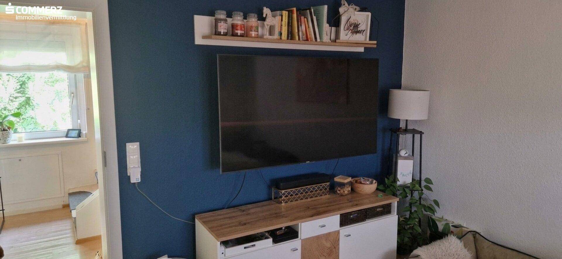 Wohnzimmer mit blau akzentuierter Wand, großem Fernseher und Fenster mit Blick ins Grüne.