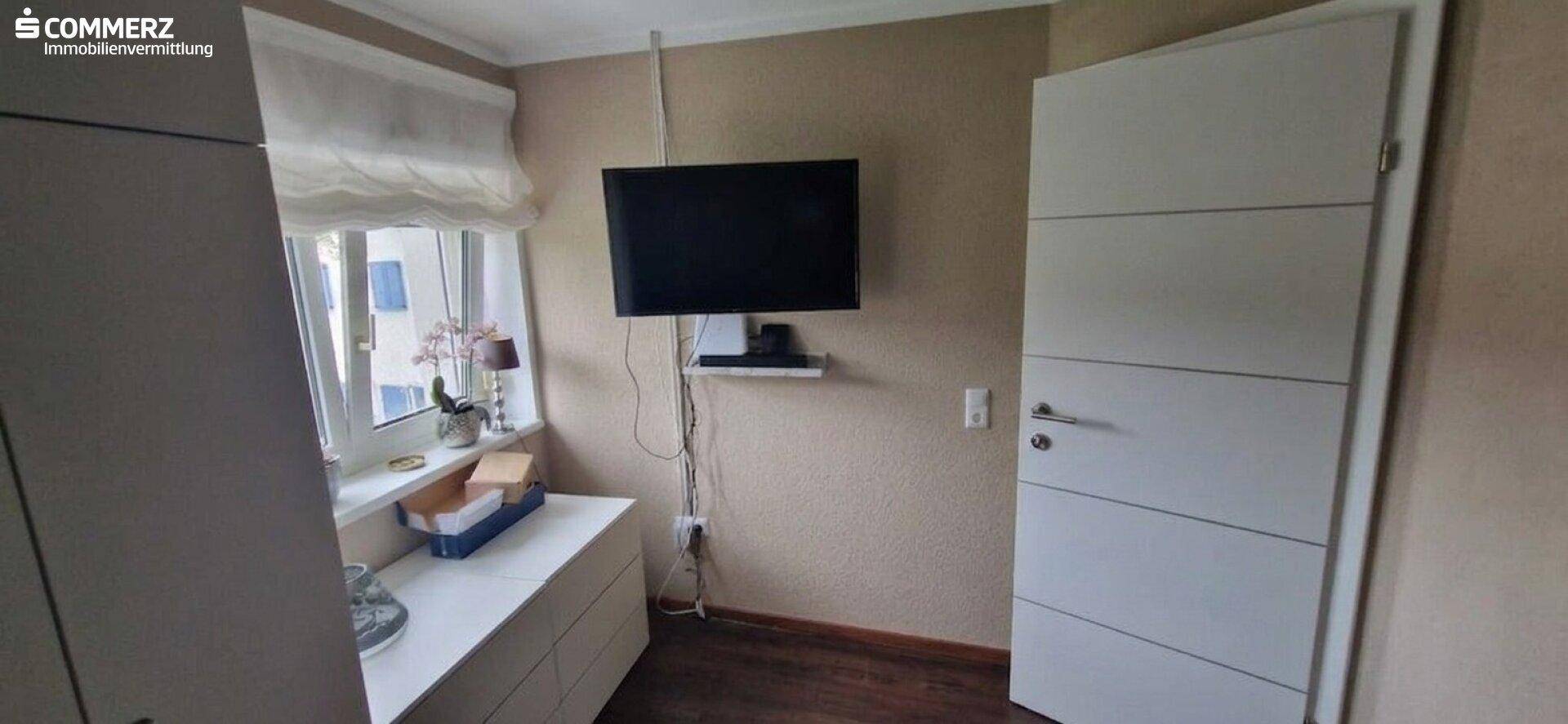 Schlafzimmer mit Fernseher an der Wand, Fenster und schlichten Möbeln.