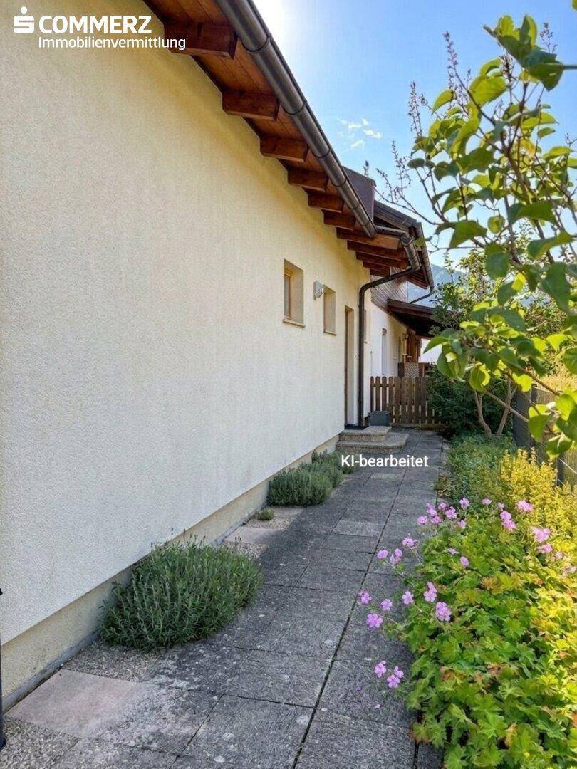 Seitenansicht des Hauses mit gepflastertem Weg, Blumenbeeten und schlichter Fassadengestaltung.