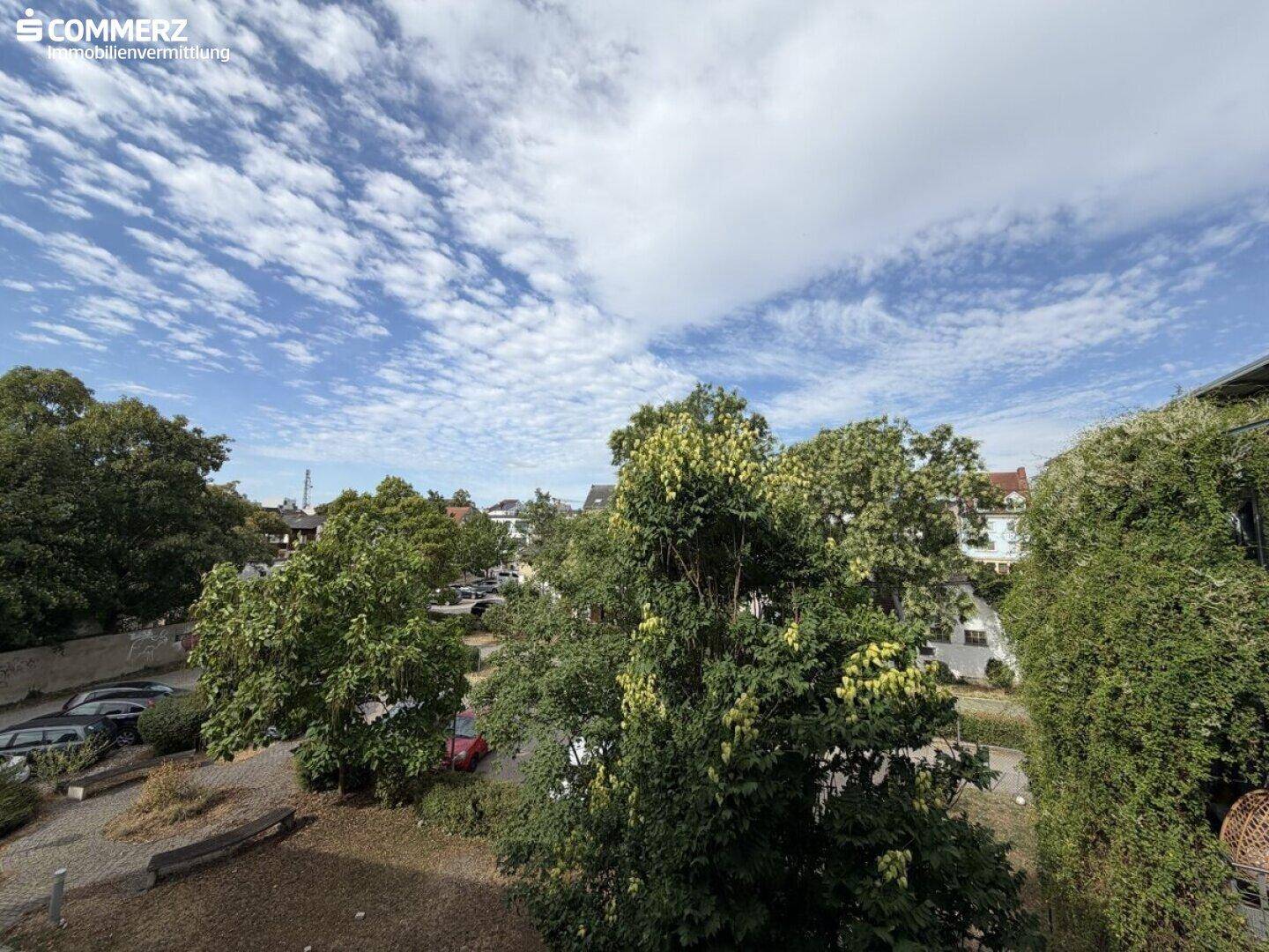 Ausblick vom Balkon auf grüne Bäume, einen Parkplatz und den Himmel.