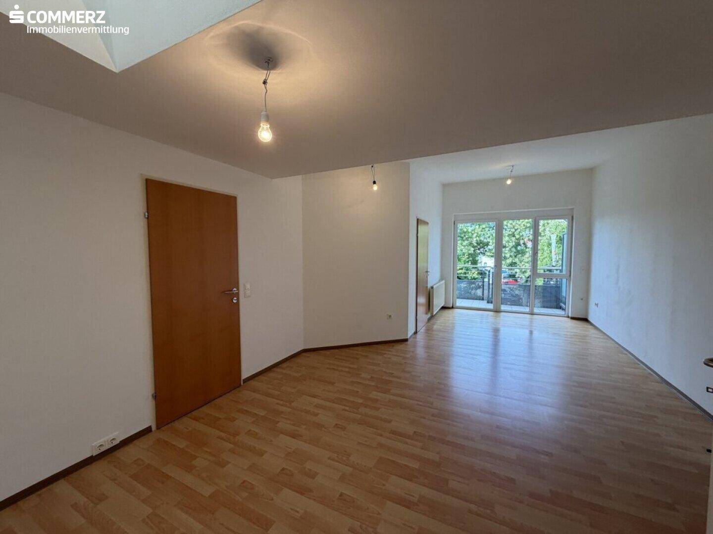 Helles, leeres Wohnzimmer mit Laminatboden und Zugang zu einem Balkon.