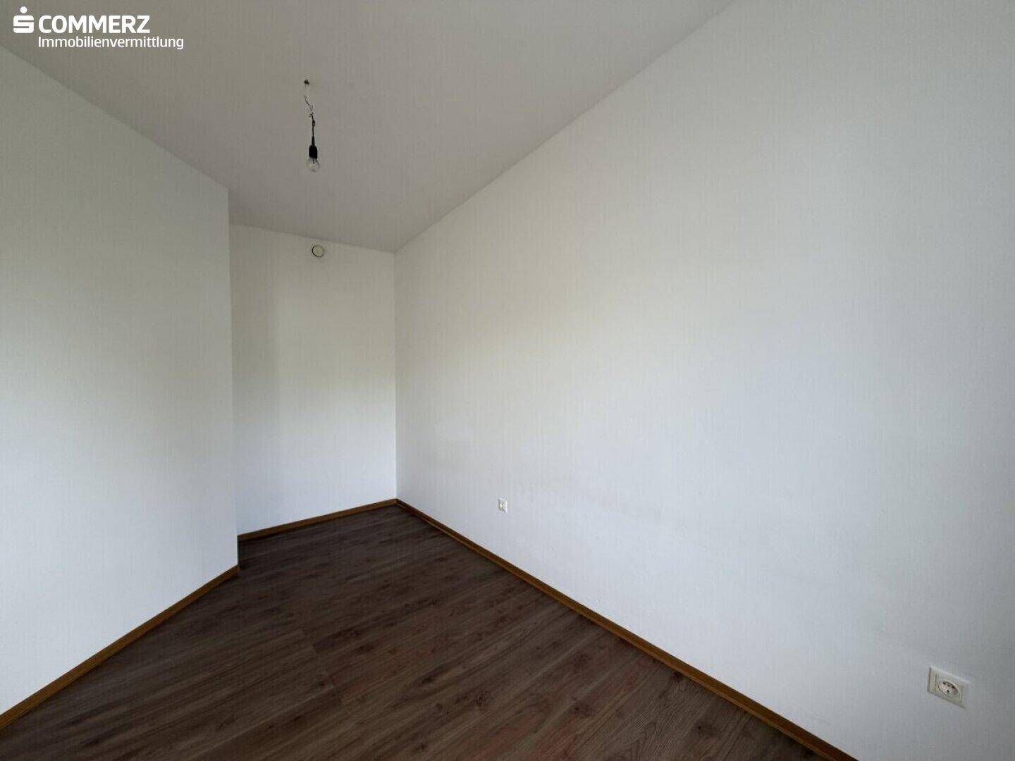 Unmöbliertes Zimmer mit dunklem Laminatboden und schlichten weißen Wänden.