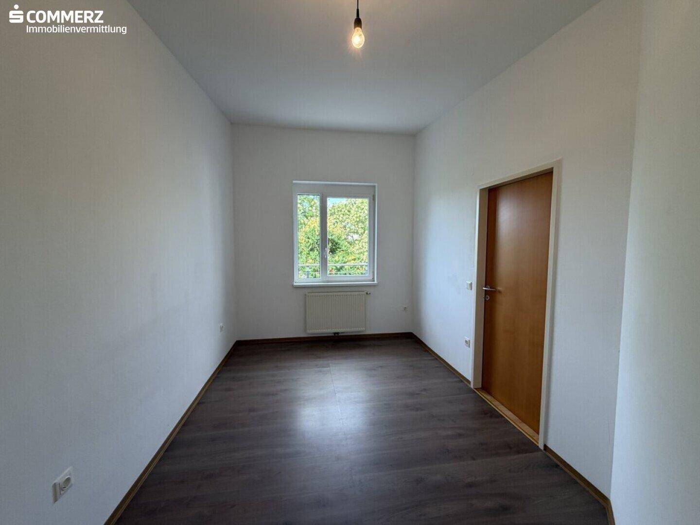 Leeres Zimmer mit dunklem Laminatboden, weiß gestrichenen Wänden und Fensterblick ins Grüne.