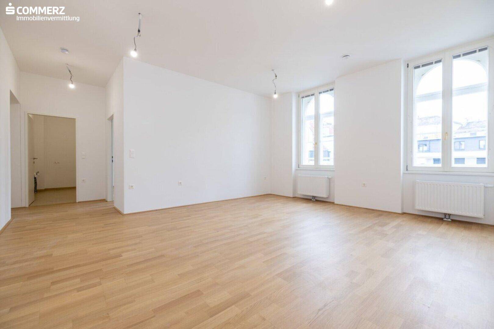 Geräumiger, heller Raum mit Parkettboden und hohen Fenstern, ideal als Wohnzimmer.