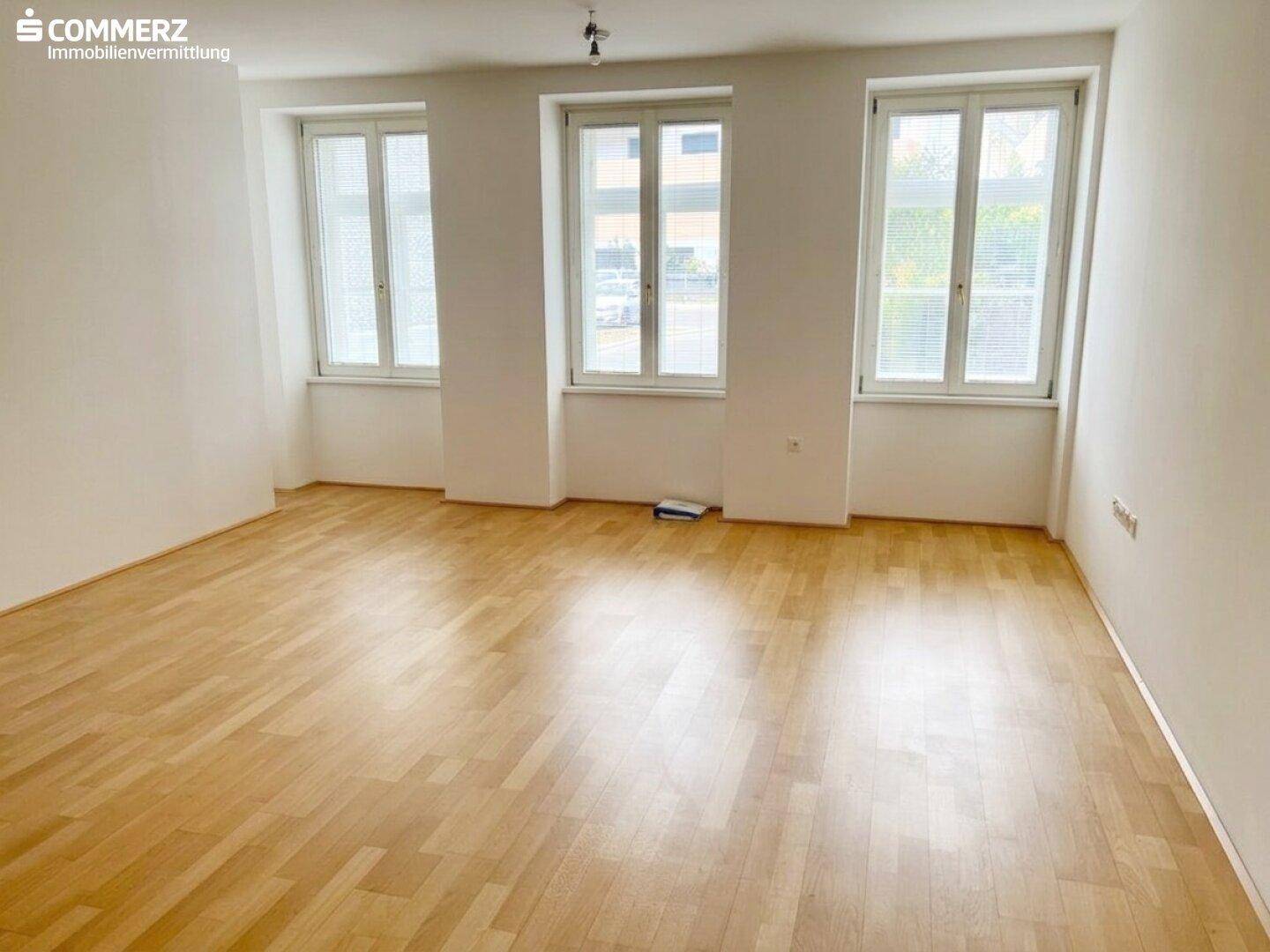 Geräumiger Wohnbereich mit hellem Parkett und hohen Fenstern, die den Raum lichtdurchfluten.