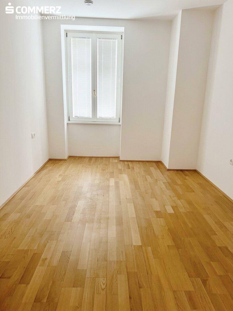 Helles Schlafzimmer mit Parkettboden und einem großen Fenster, ideal für Ruhe und Entspannung.