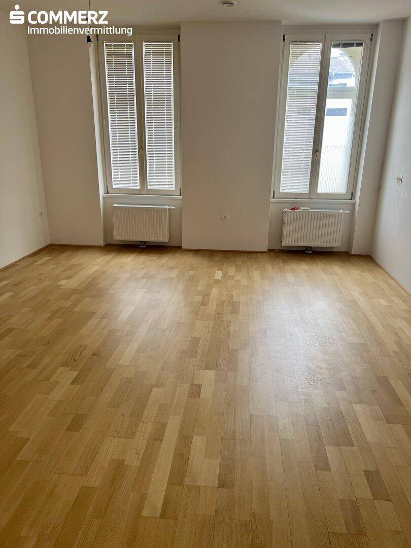 Leerer Raum mit Holzboden und zwei hohen Fenstern, bietet viel natürliches Licht.