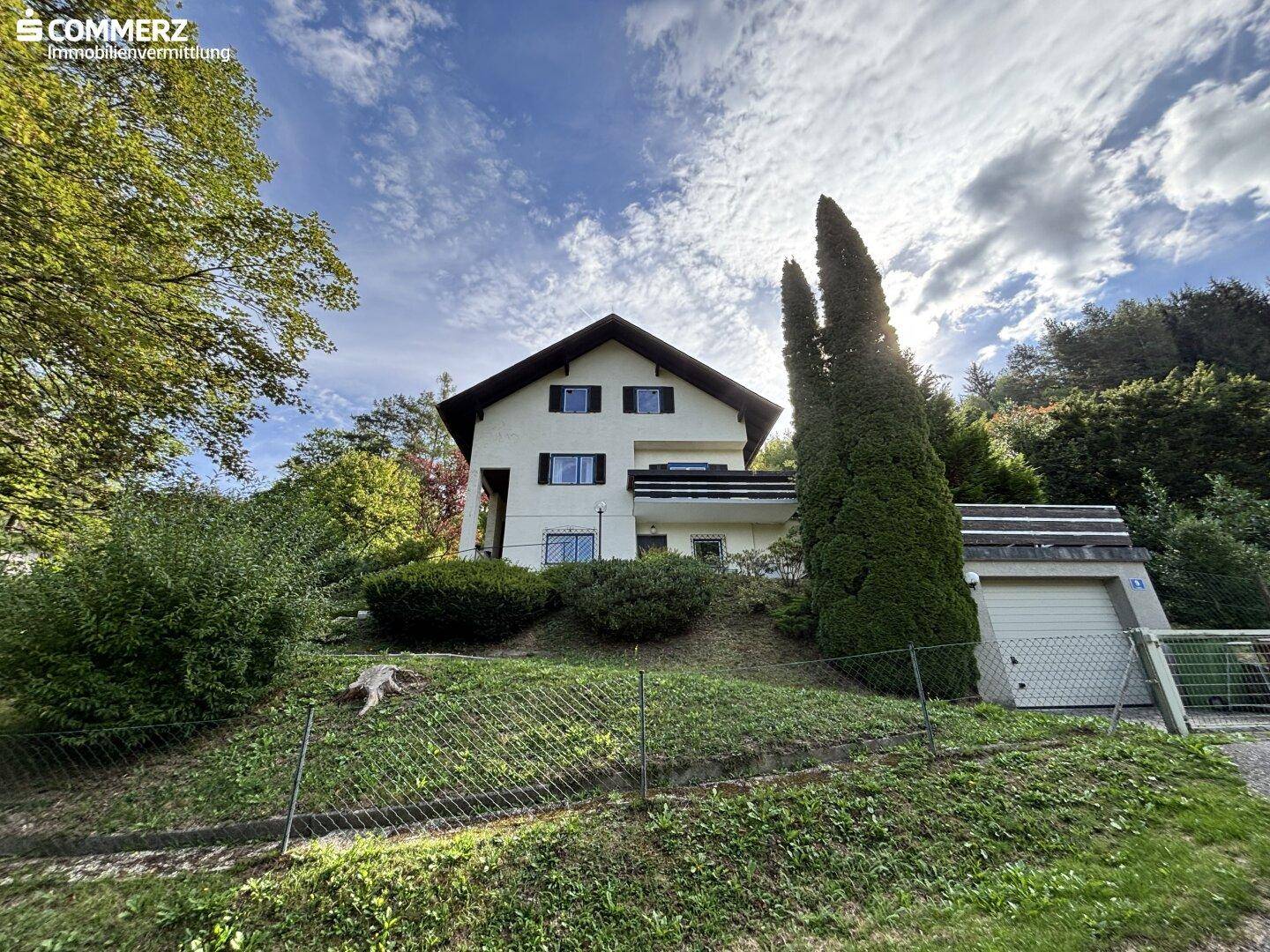 Haus mit Garage und Balkon, eingebettet in eine grüne Hanglage mit hohen Bäumen.