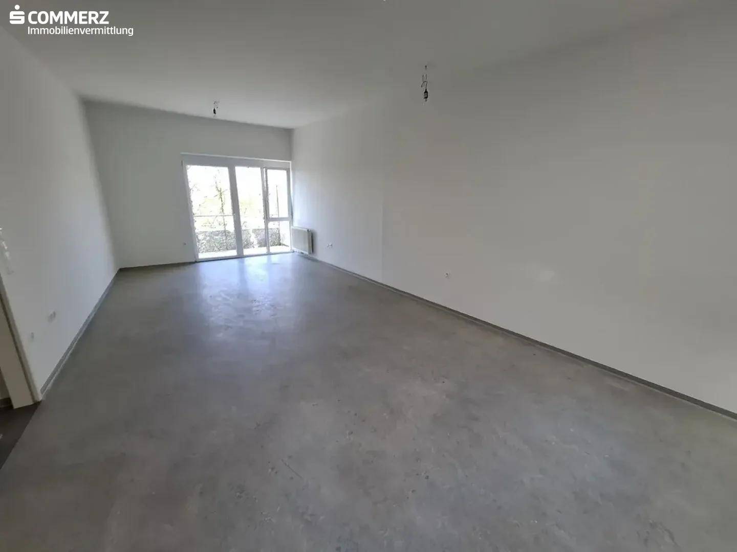 Geräumiges, leeres Wohnzimmer mit hellen Wänden, Betonboden und Zugang zu einem Balkon.