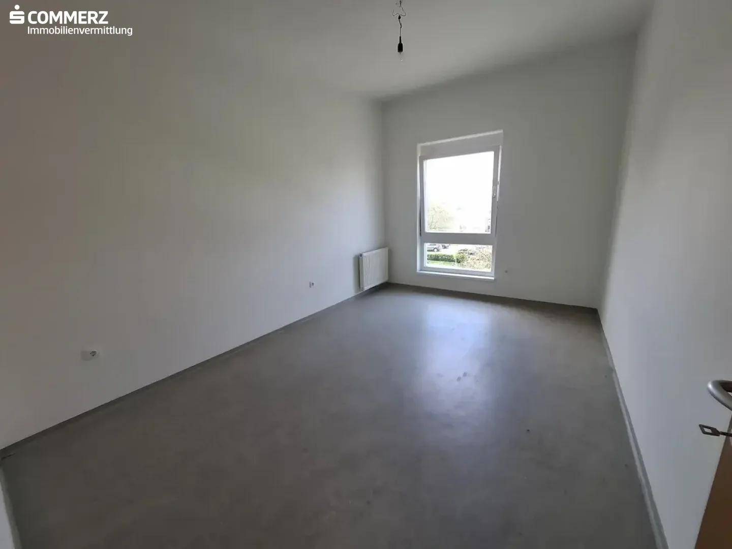 Leeres Schlafzimmer mit hellen Wänden, Betonboden und einem Fenster mit Blick ins Grüne.