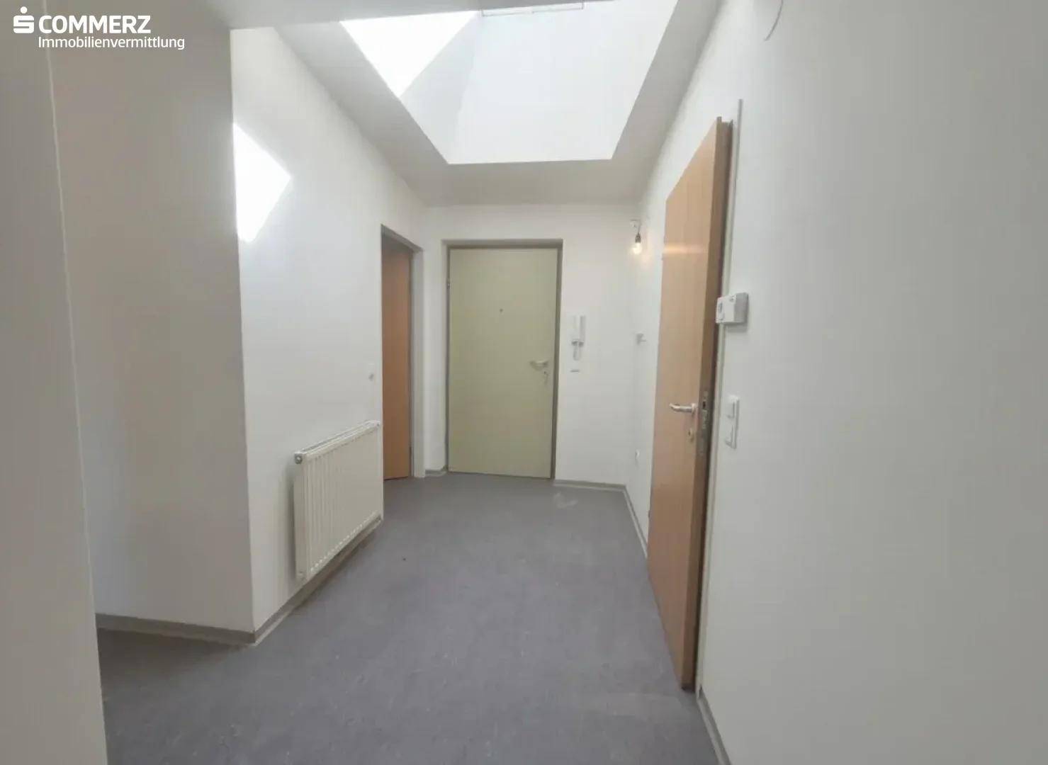 Heller Flur mit Betonboden, mehreren Türen und einem Oberlicht für natürliches Licht.