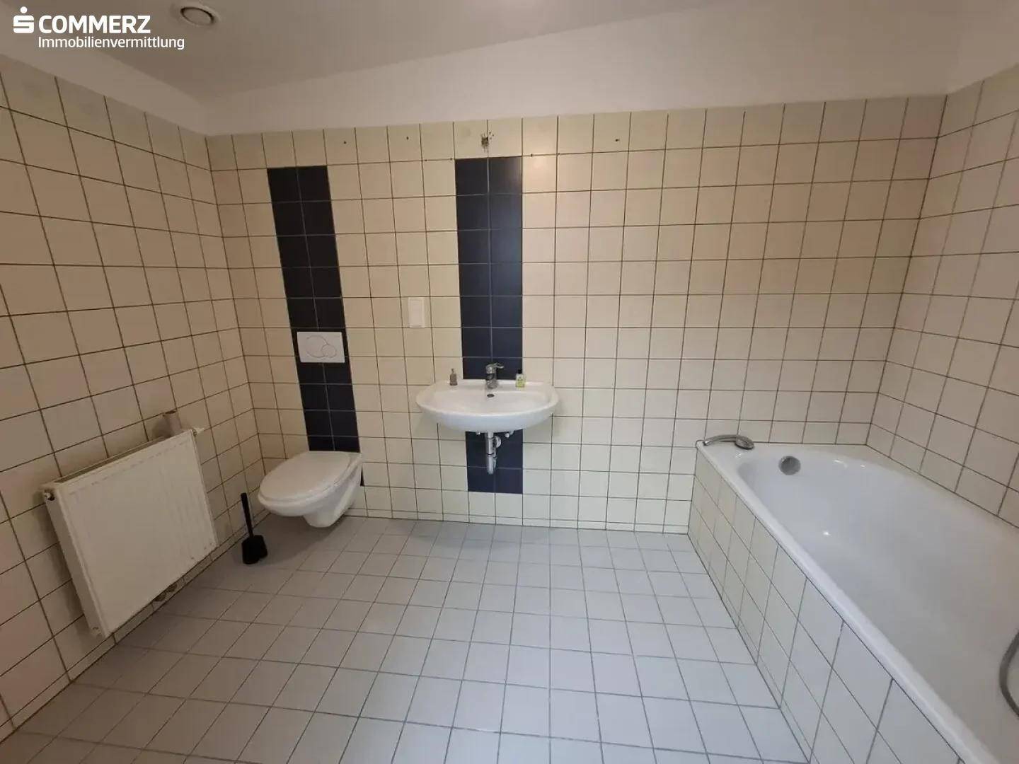 Badezimmer mit Badewanne, WC, Waschbecken und hellen Fliesen mit dekorativem Streifen.