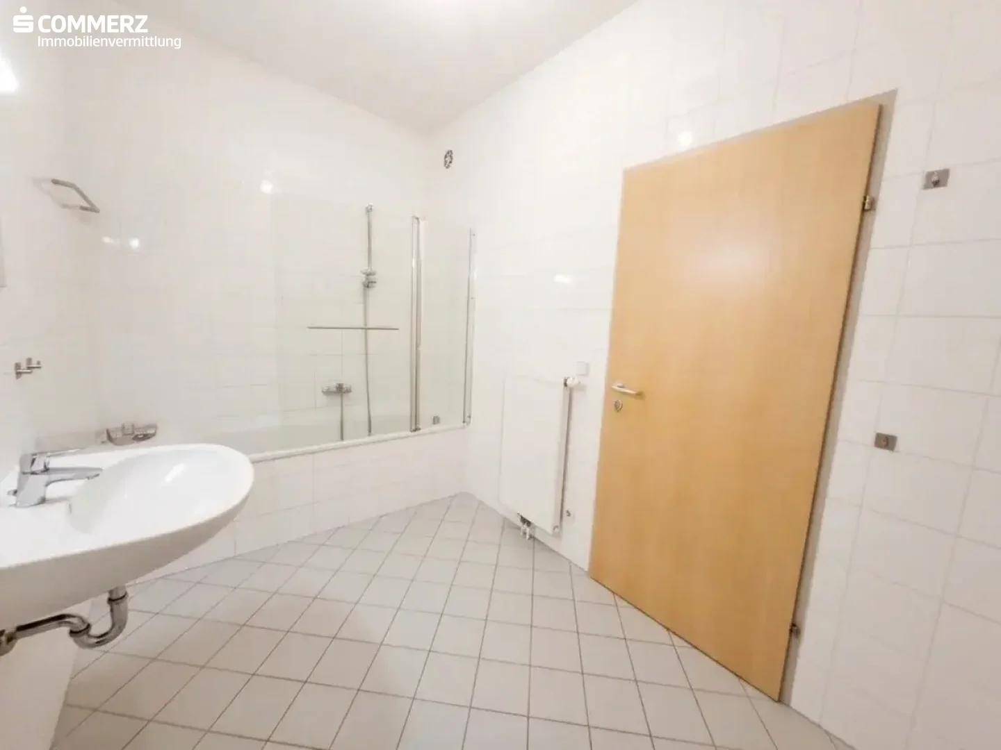 Helles Badezimmer mit weißen Fliesen, Badewanne mit Duschfunktion und Waschbecken.