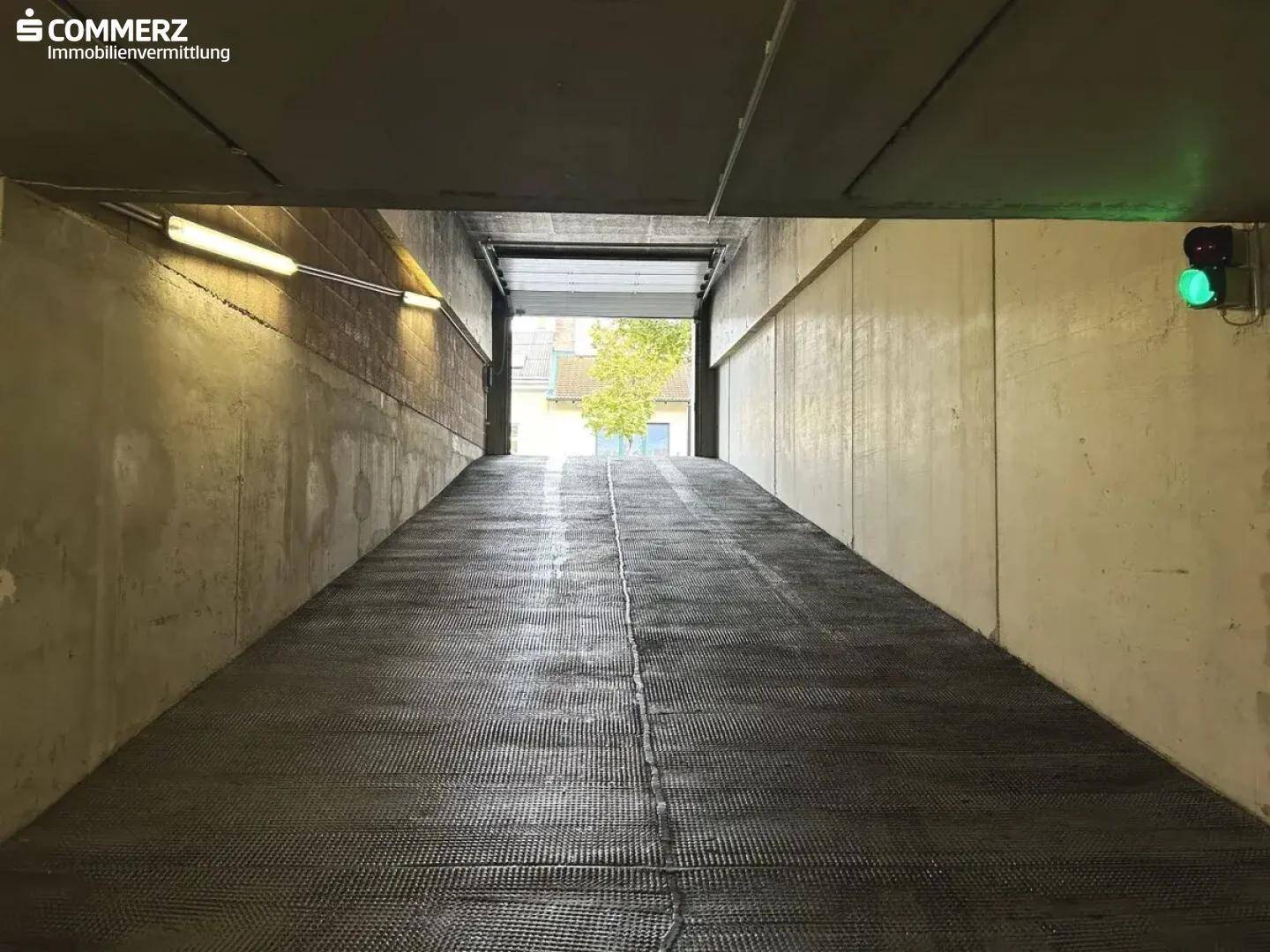 Ausfahrt einer Tiefgarage mit Rampe und grünem Ampelsignal, führt ins Freie mit Blick auf Bäume.