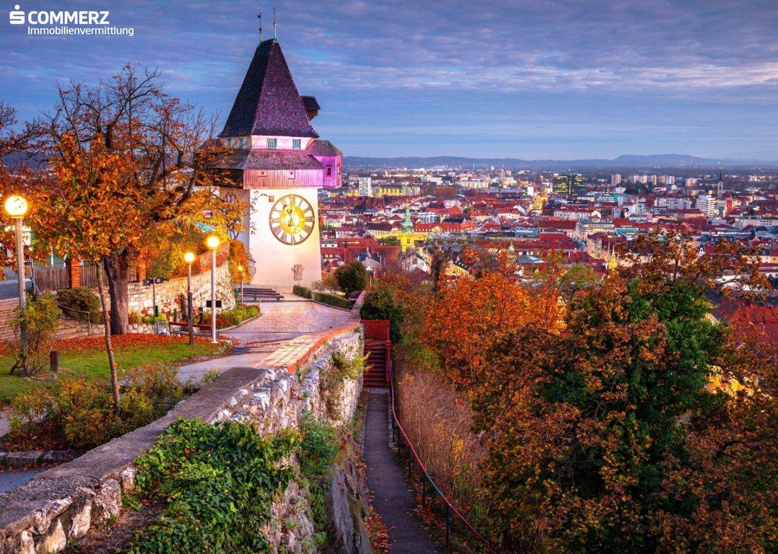 Historischer Uhrturm von Graz bei Dämmerung mit herbstlicher Stadtkulisse und beleuchteten Wegen.