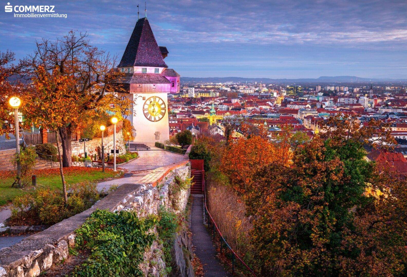 Panoramablick auf die Stadt bei Dämmerung mit dem historischen Uhrturm und herbstlicher Vegetation.