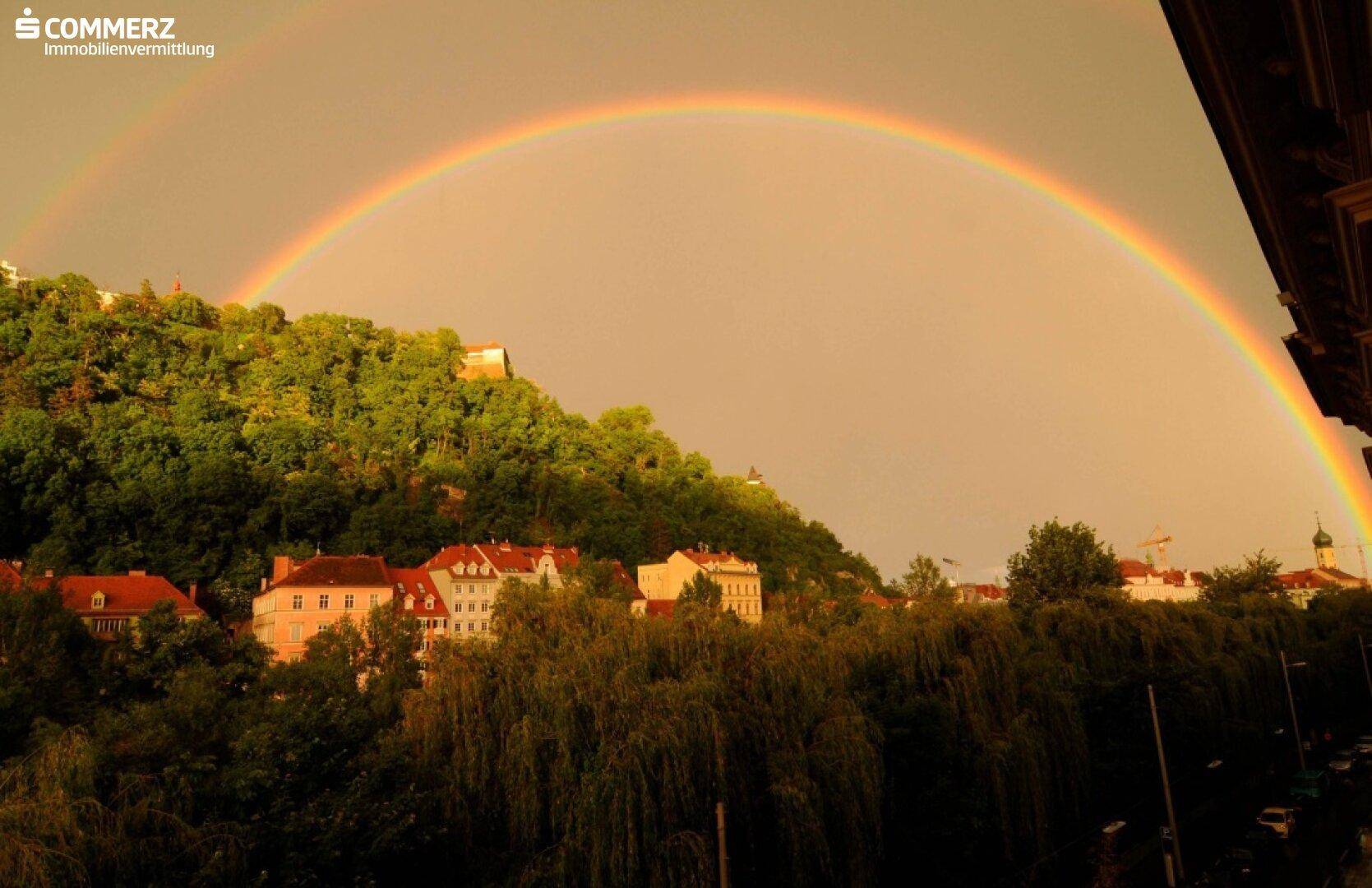 Doppelter Regenbogen über einer grünen Hügellandschaft mit Häusern und Bäumen.