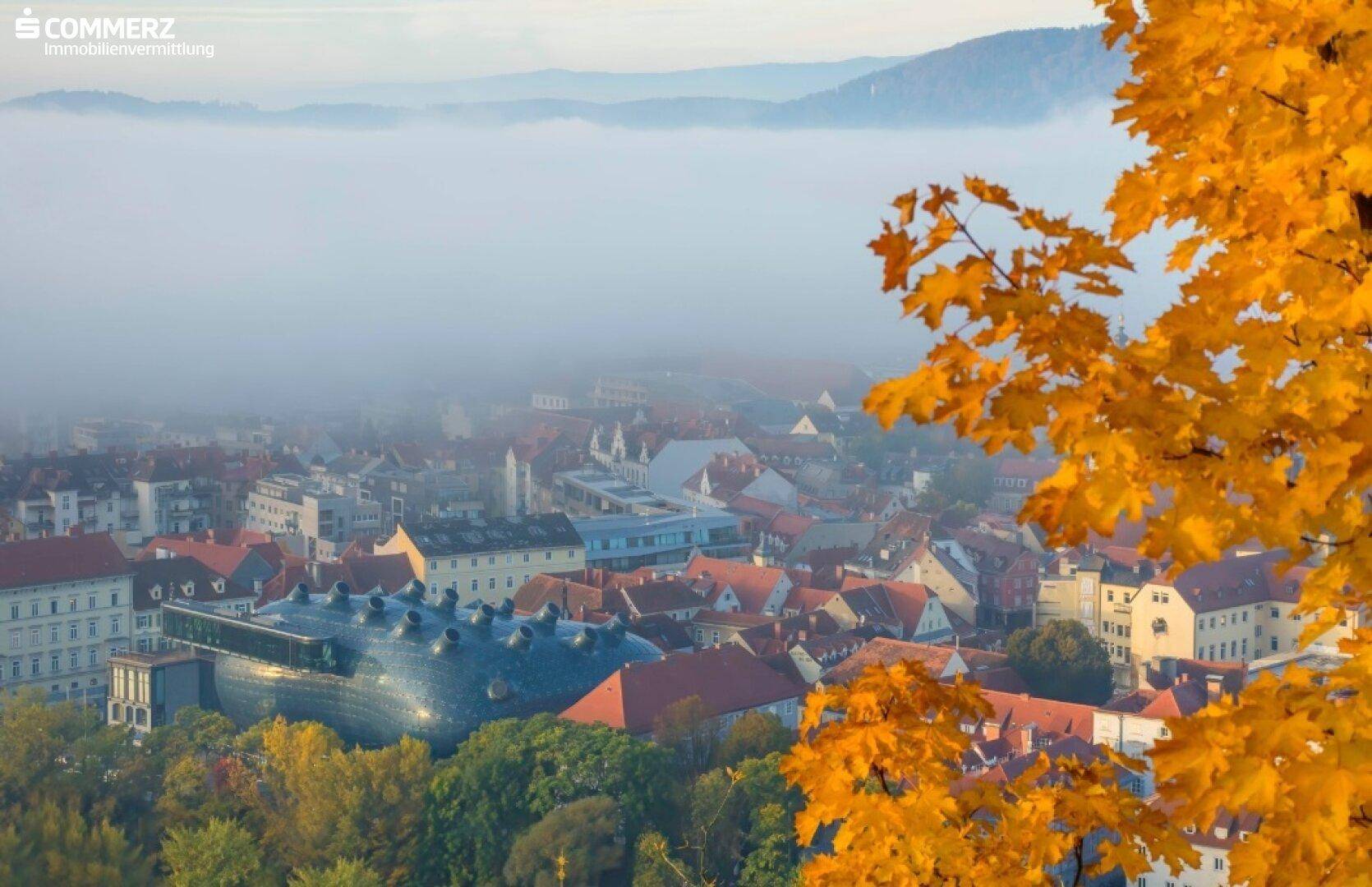 Nebelverhangene Stadtlandschaft mit herbstlichen Bäumen im Vordergrund und Bergen im Hintergrund.