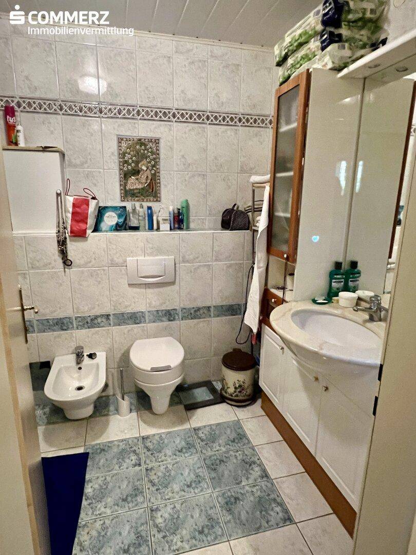 Badezimmer mit hellblauen und weißen Fliesen, ausgestattet mit Toilette, Bidet und Waschbecken.