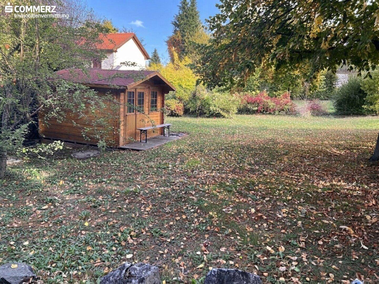 Großer Gartenbereich mit Holzschuppen, Rasenfläche und herbstlichem Laub, umgeben von Bäumen.