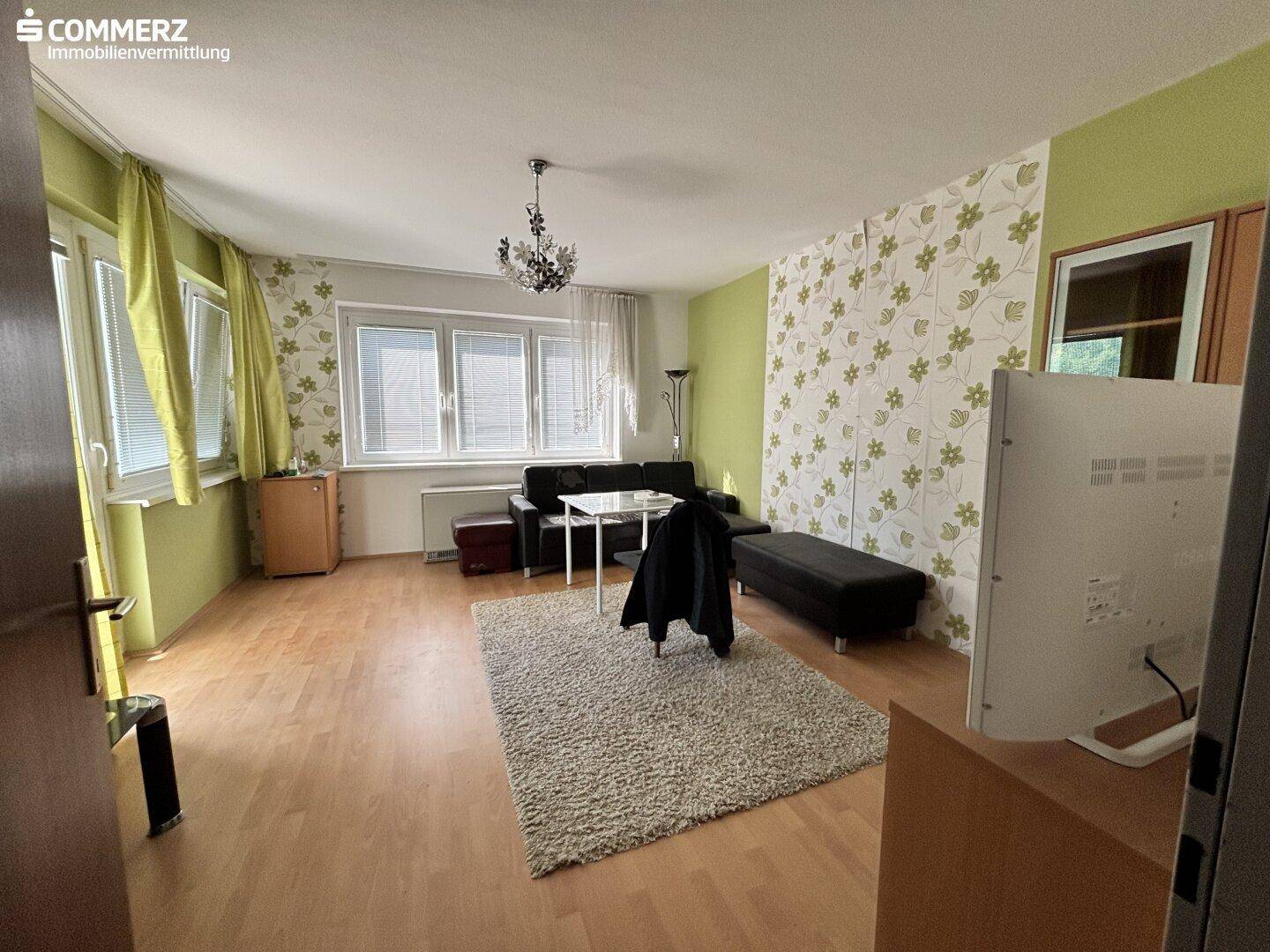 Helles Wohnzimmer mit Parkettboden, floraler Tapete und einem Sofa unter großen Fenstern.