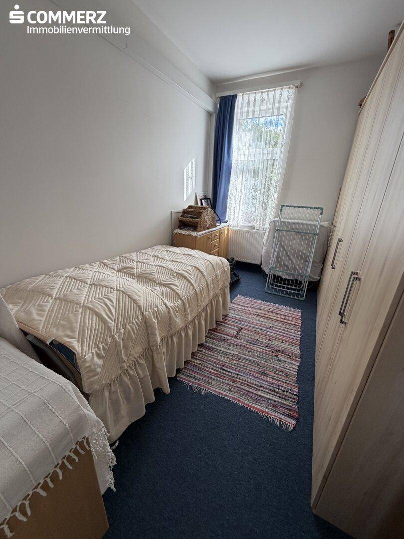 Schlafzimmer mit Einzelbett, Kleiderschrank und Teppichboden, hell durch Fenster.