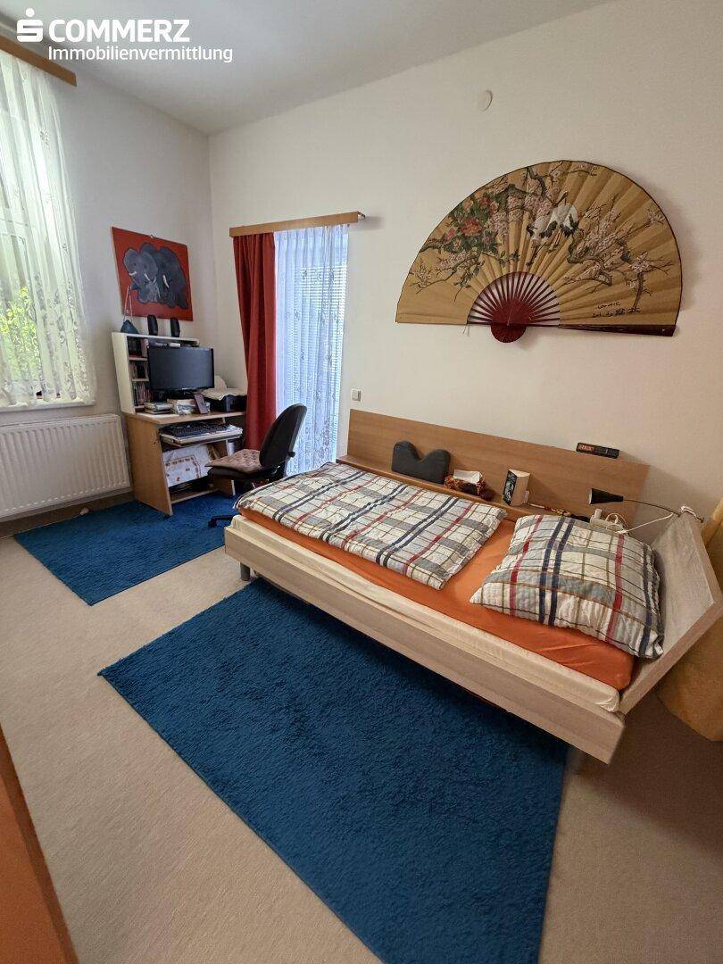Schlafzimmer mit Bett, Schreibtisch und dekorativem Fächer an der Wand.