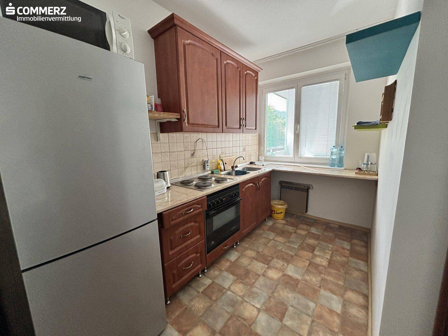 Küche mit braunen Holzschränken, gefliestem Boden und einem Kühlschrank neben dem Fenster.