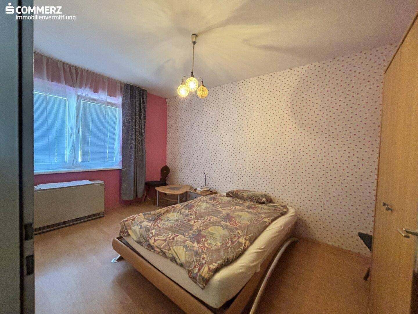 Schlafzimmer mit Parkettboden, gemusterter Tapete und einem Doppelbett unter einem Fenster.