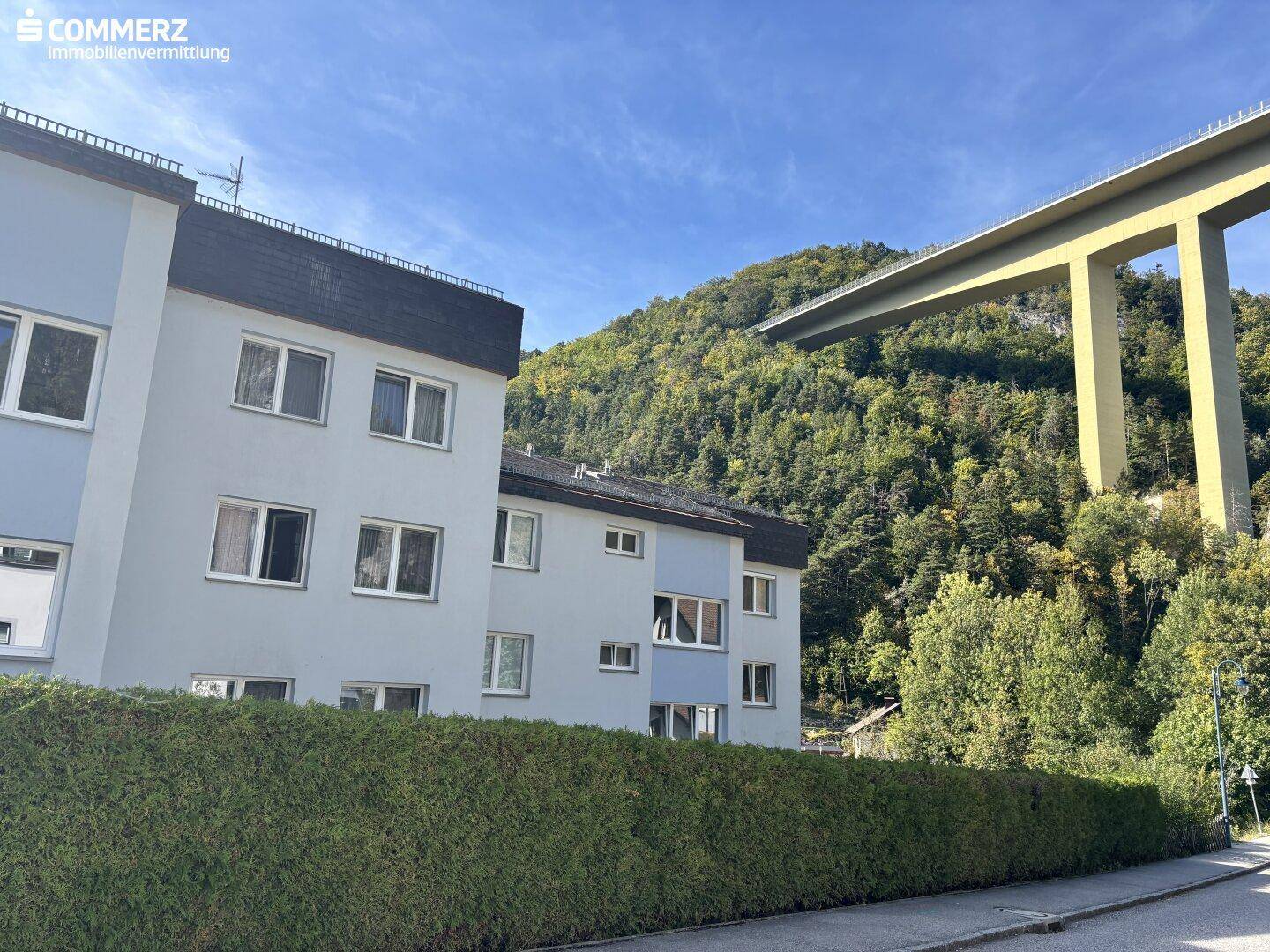 Mehrfamilienhaus mit blauer Fassade und einer hohen Hecke im Vordergrund, umgeben von grünen Hügeln.