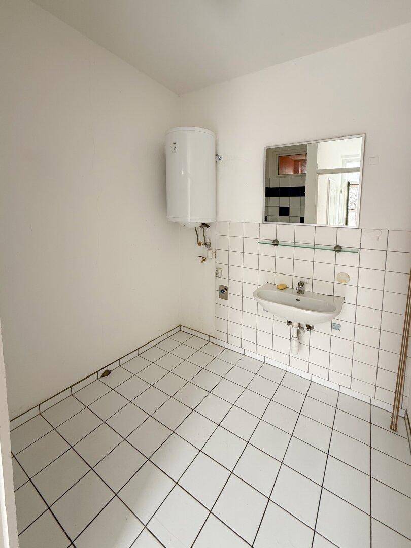 Nutzraum mit Waschbecken, Warmwasserboiler und weißen Fliesen an Wänden und Boden.