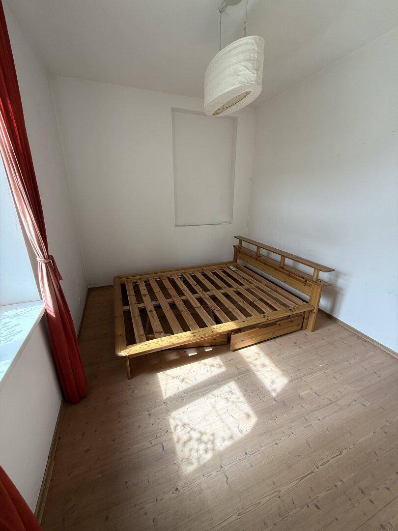 Schlafzimmer mit Holzdielenboden, einem Holzrahmenbett und einem Fenster mit rotem Vorhang.