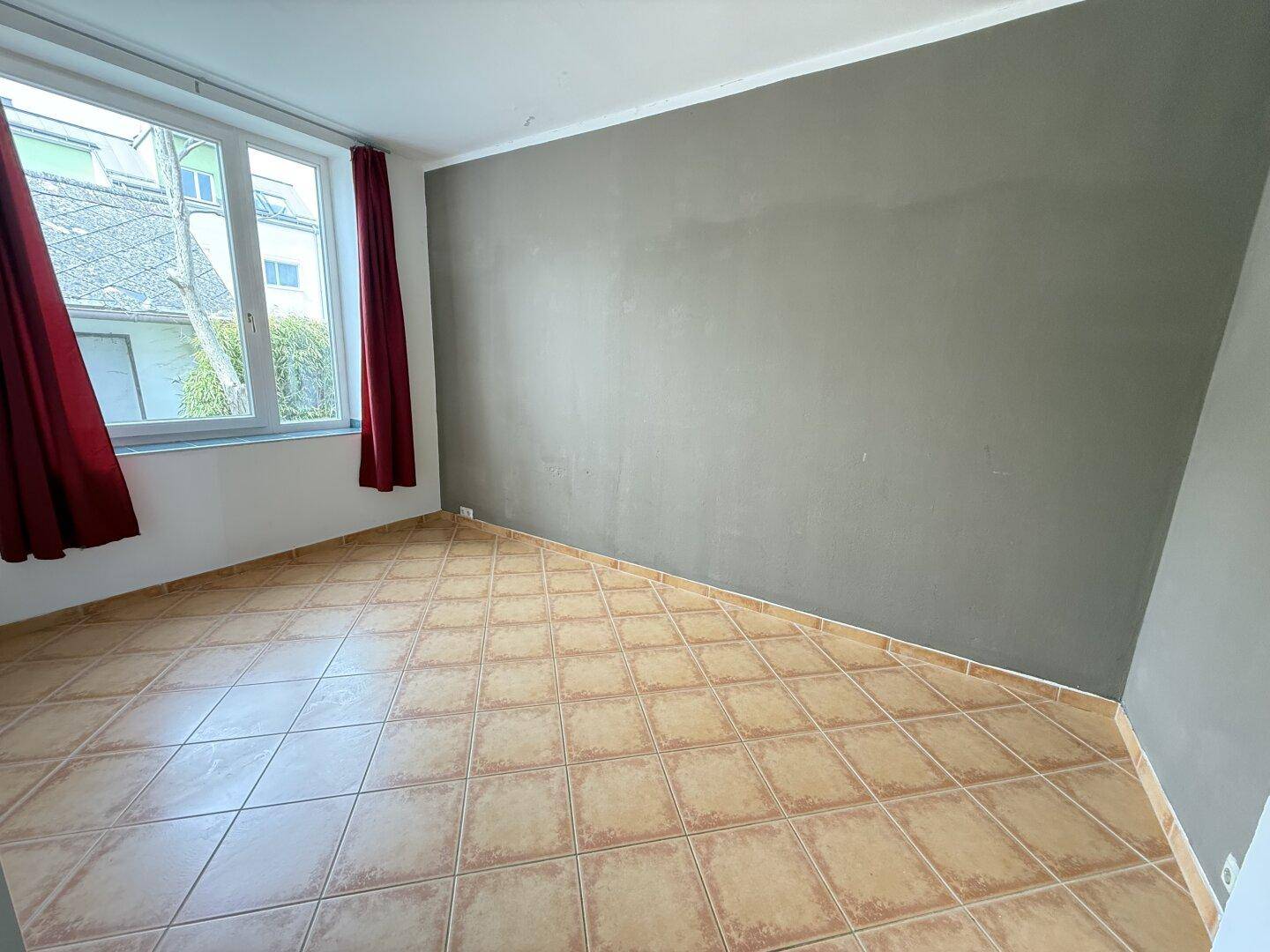 Geräumiges Zimmer mit Fliesenboden, einer grauen Akzentwand und großen Fenstern mit roten Vorhängen.