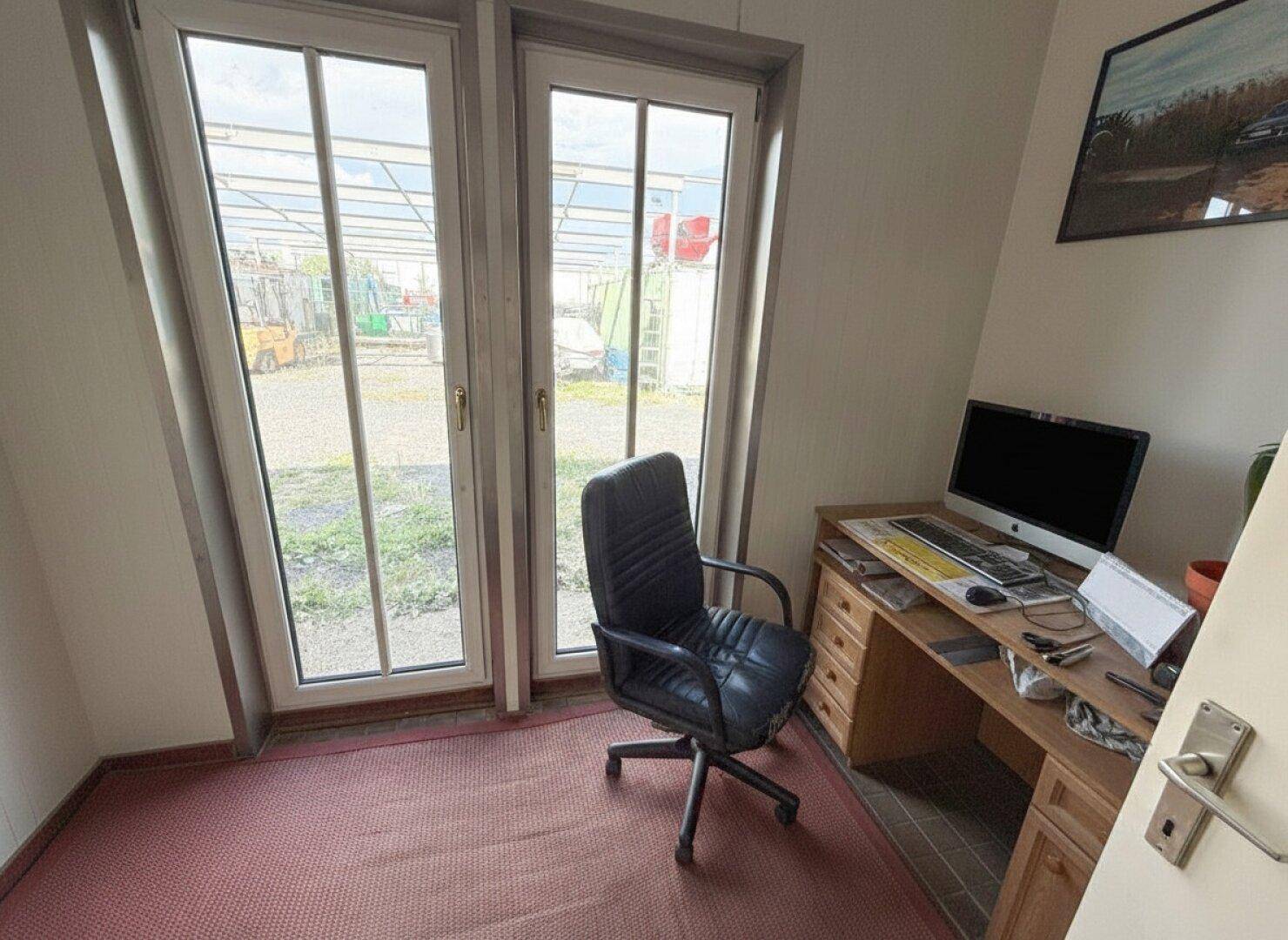 Kleines Büro mit einem Holzschreibtisch, Bürostuhl und großen Fenstertüren, die viel Tageslicht hereinlassen.