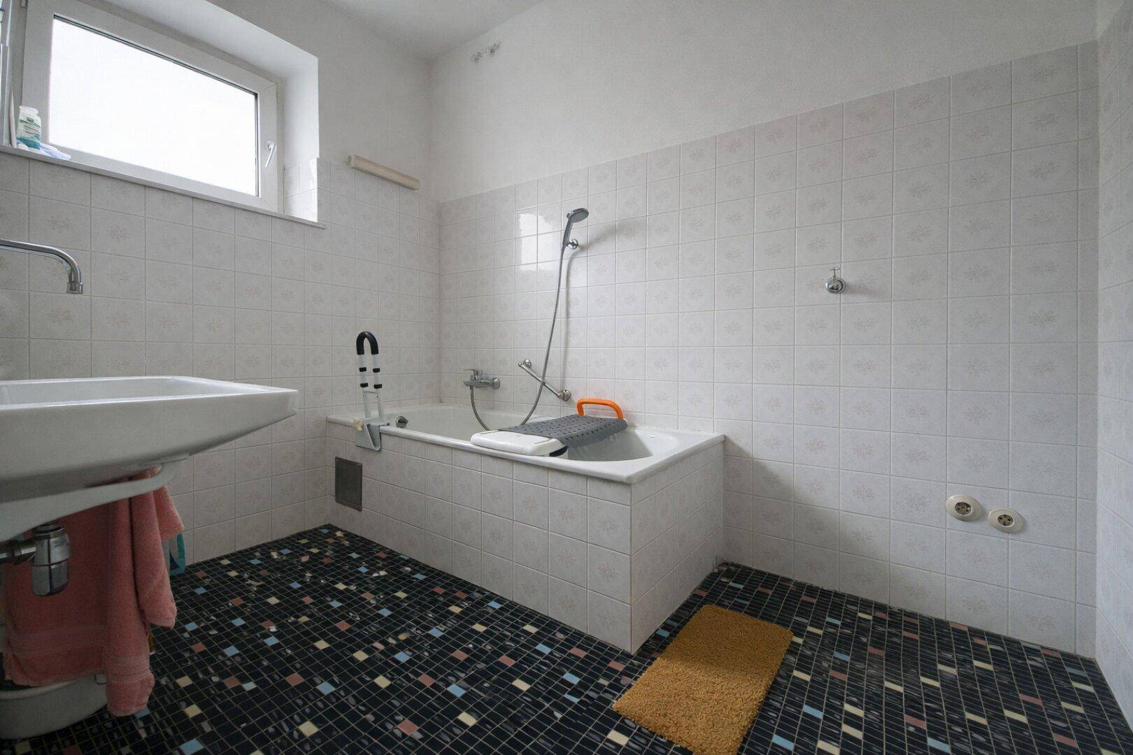 Badezimmer mit Badewanne, Waschbecken und auffälligen Bodenfliesen im 80er-Jahre-Stil.