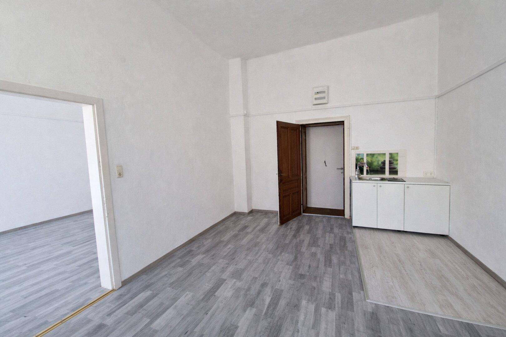 Heller Raum mit kleiner Küchenzeile, grauem Laminatboden und Zugang zu einem weiteren Zimmer.