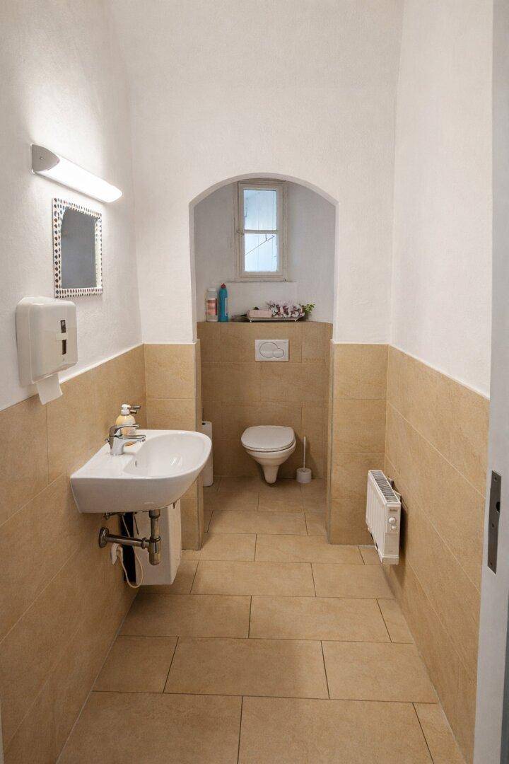 Helles Badezimmer mit beigefarbenen Fliesen, einem Waschbecken, Toilette und einem kleinen Fenster in einer Nische.