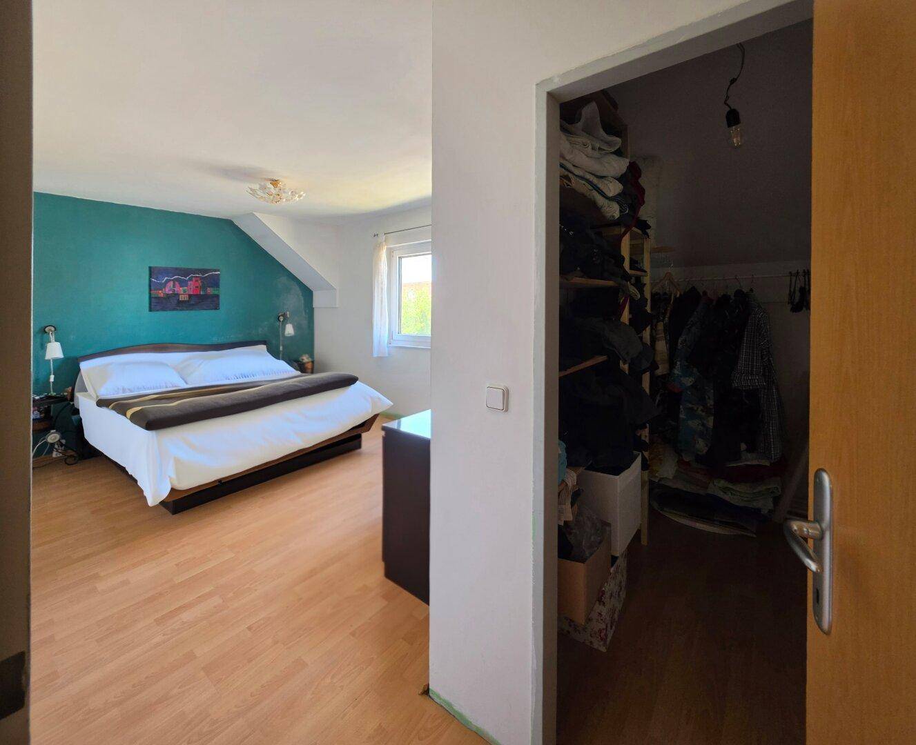 Schlafzimmer mit farbiger Akzentwand, Laminatboden und angrenzendem begehbarem Kleiderschrank.